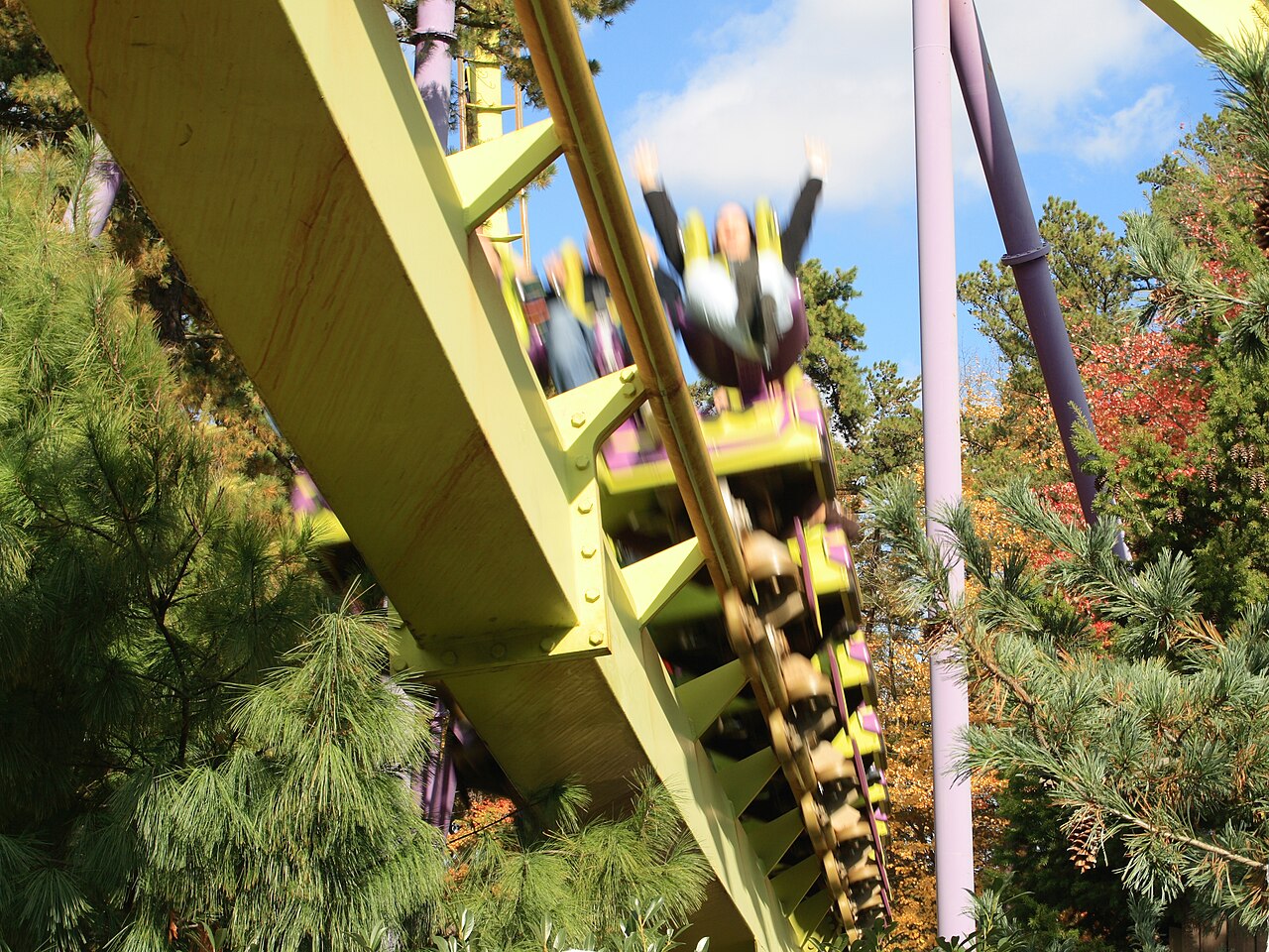 A Medusa floorless train rushes past trees close to the queue with riders' hands in the air