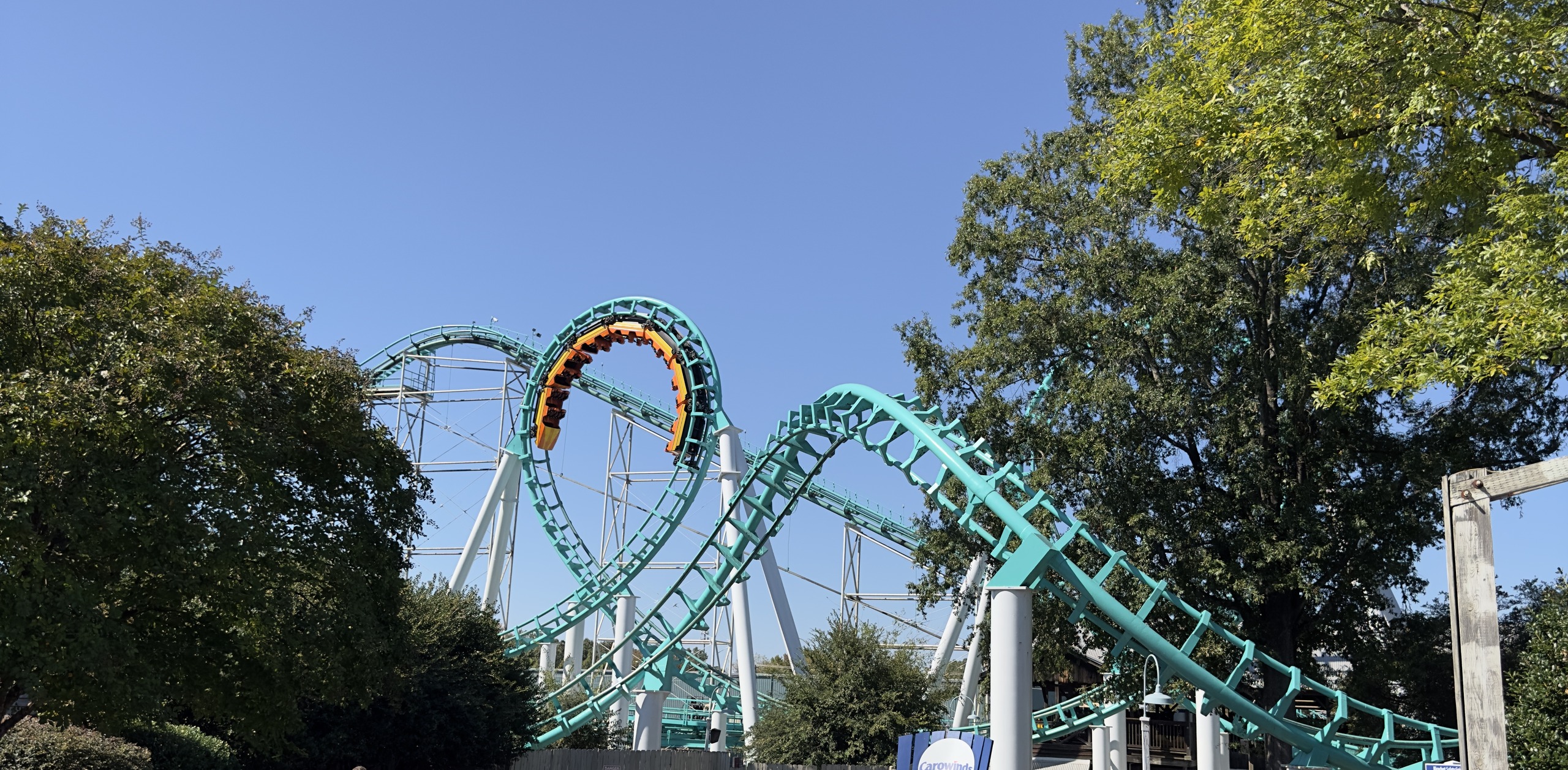 An orange Carolina Cyclone train upside down at the top of a vertical loop, framed by trees, Carowinds, October 2024