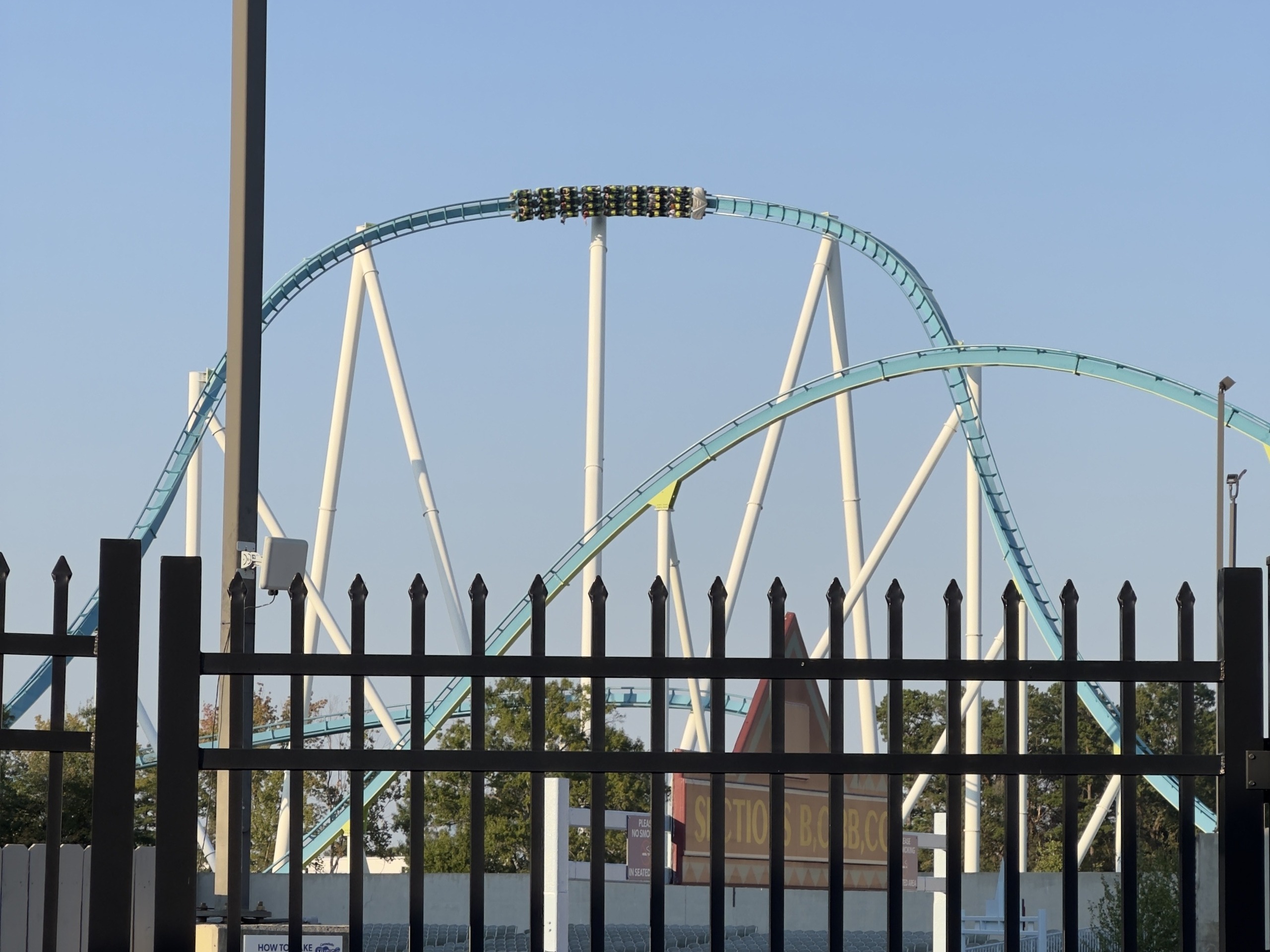 A Fury 325 train over a hill seen through the fence by the parking lot, Carowinds, October 2024