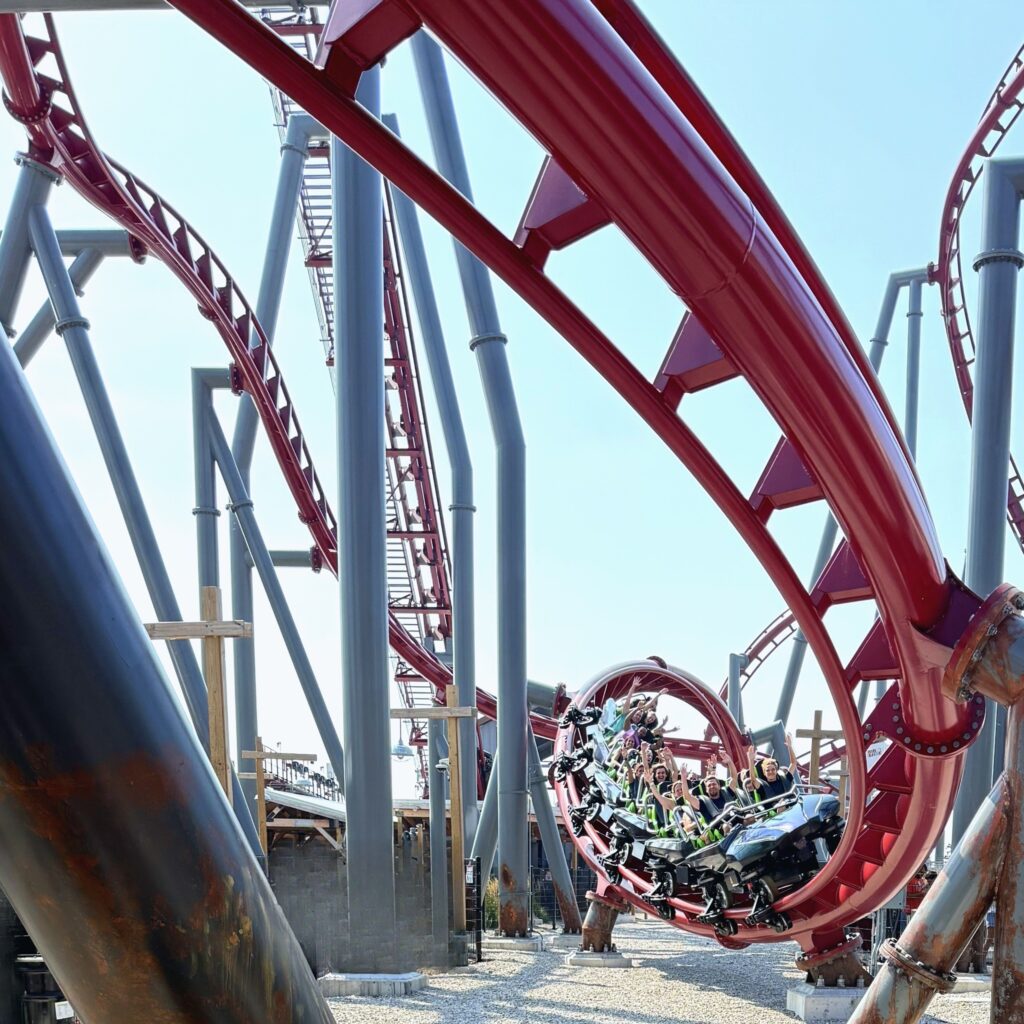 A Siren's Curse train curling through the red loop under the sweeping track at Cedar Point, seen from the gravel path below