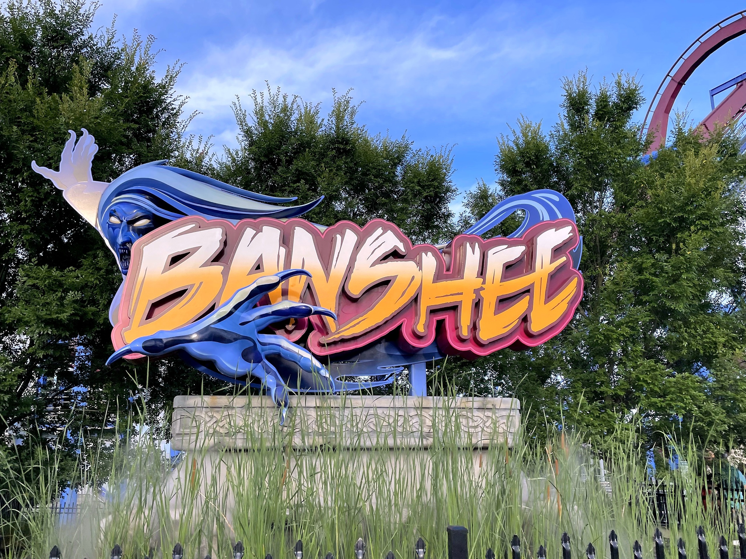 The Banshee sign and its stone base among tall ornamental grasses, Kings Island, July 2022