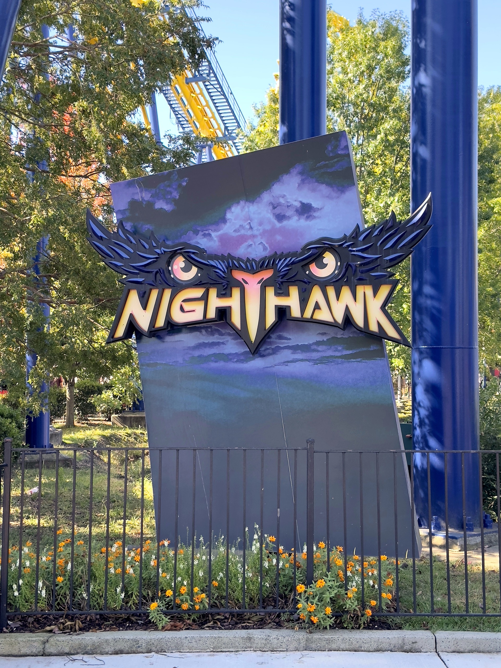 The Nighthawk entrance sign with glowing eyes in front of the blue supports and flowers at Carowinds, October 2024