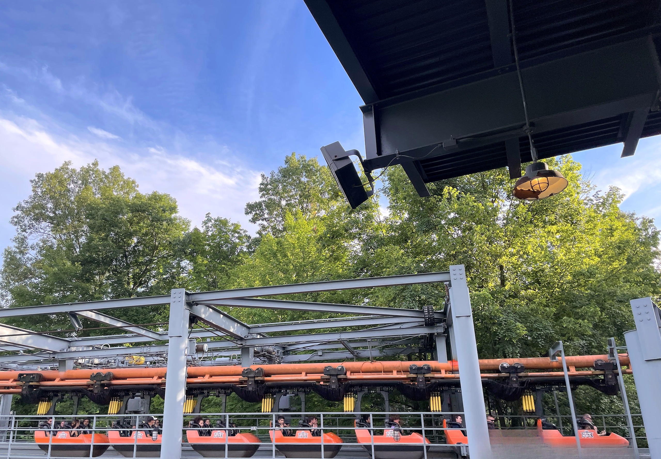 A train of orange suspended cars full of riders gliding through The Bat's station, Kings Island, July 2022