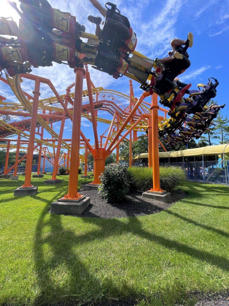 Riders hanging from a Woodstock's Air Rail train as it swings overhead through the orange supports and yellow track, Kings Island, July 2022